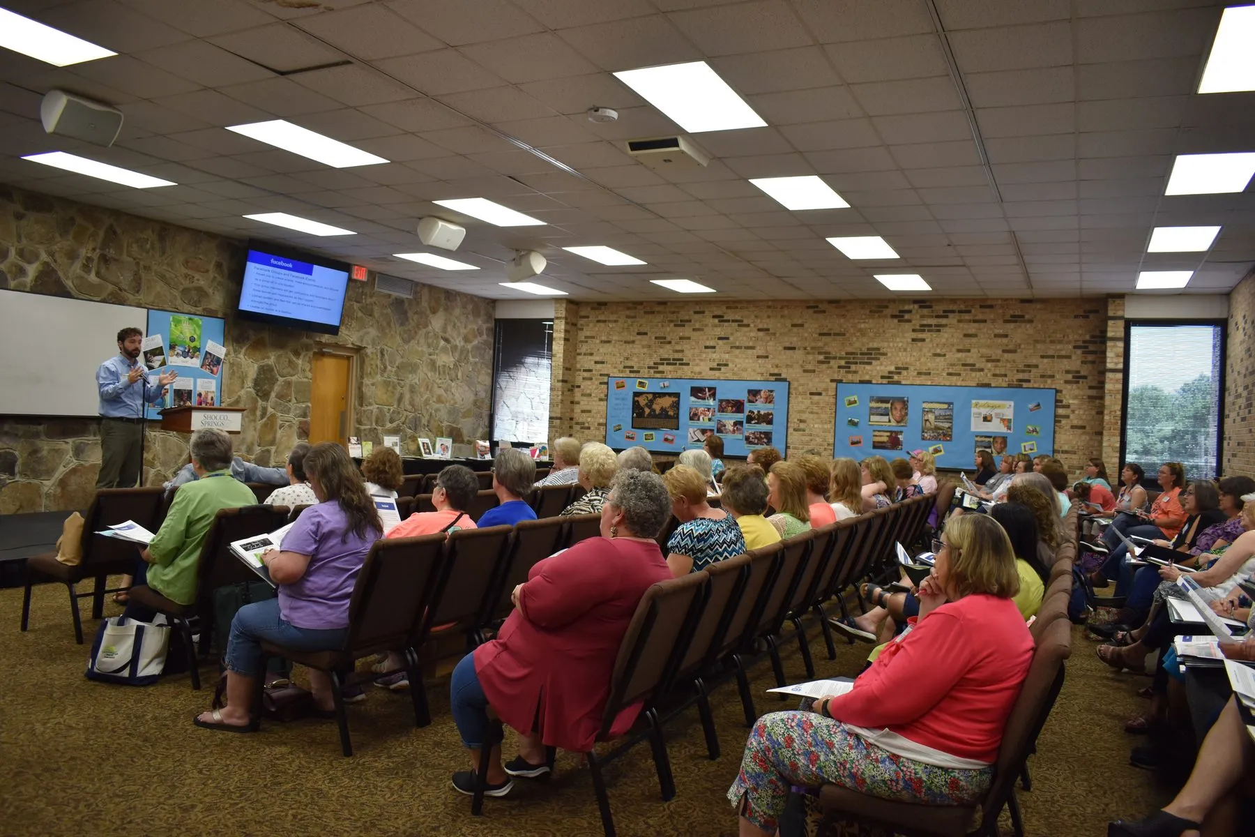 An audience of women seated at tables in a meeting room during a presentation.