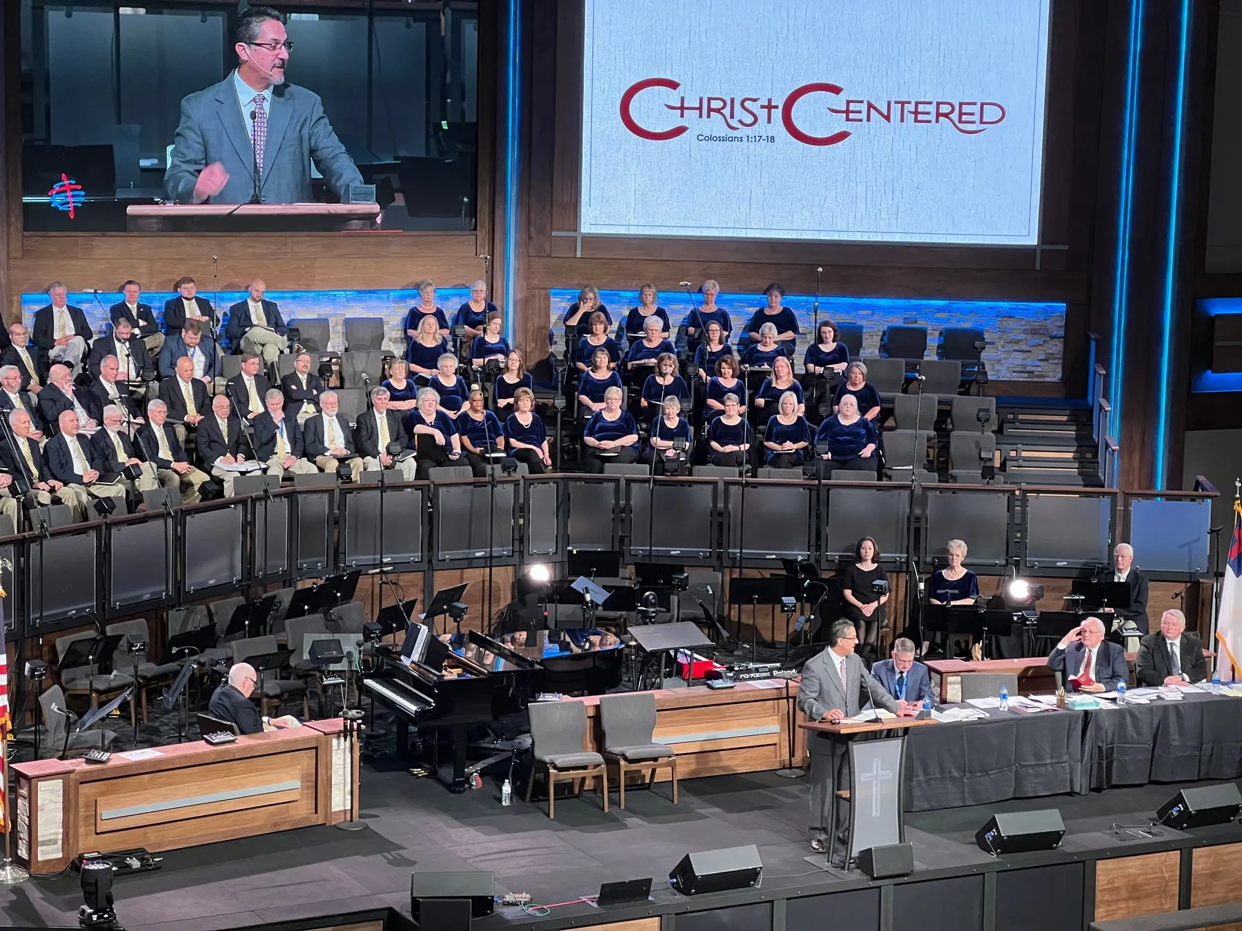 A speaker at the podium of a large convention hall, with a choir and a screen reading Christ Centered behind him.